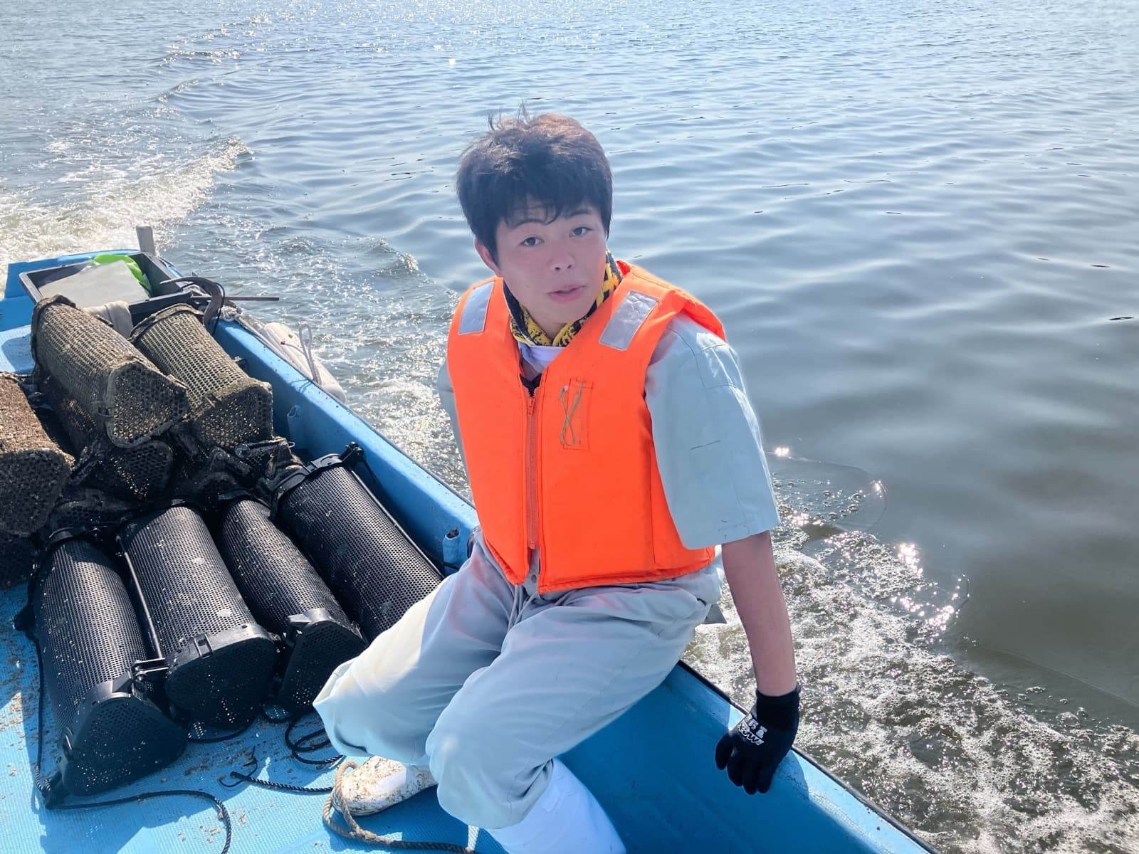 ライフジャケットを着て船の上に座る本人。傍らに牡蠣養殖のカゴが積まれ、まわりには海が広がる