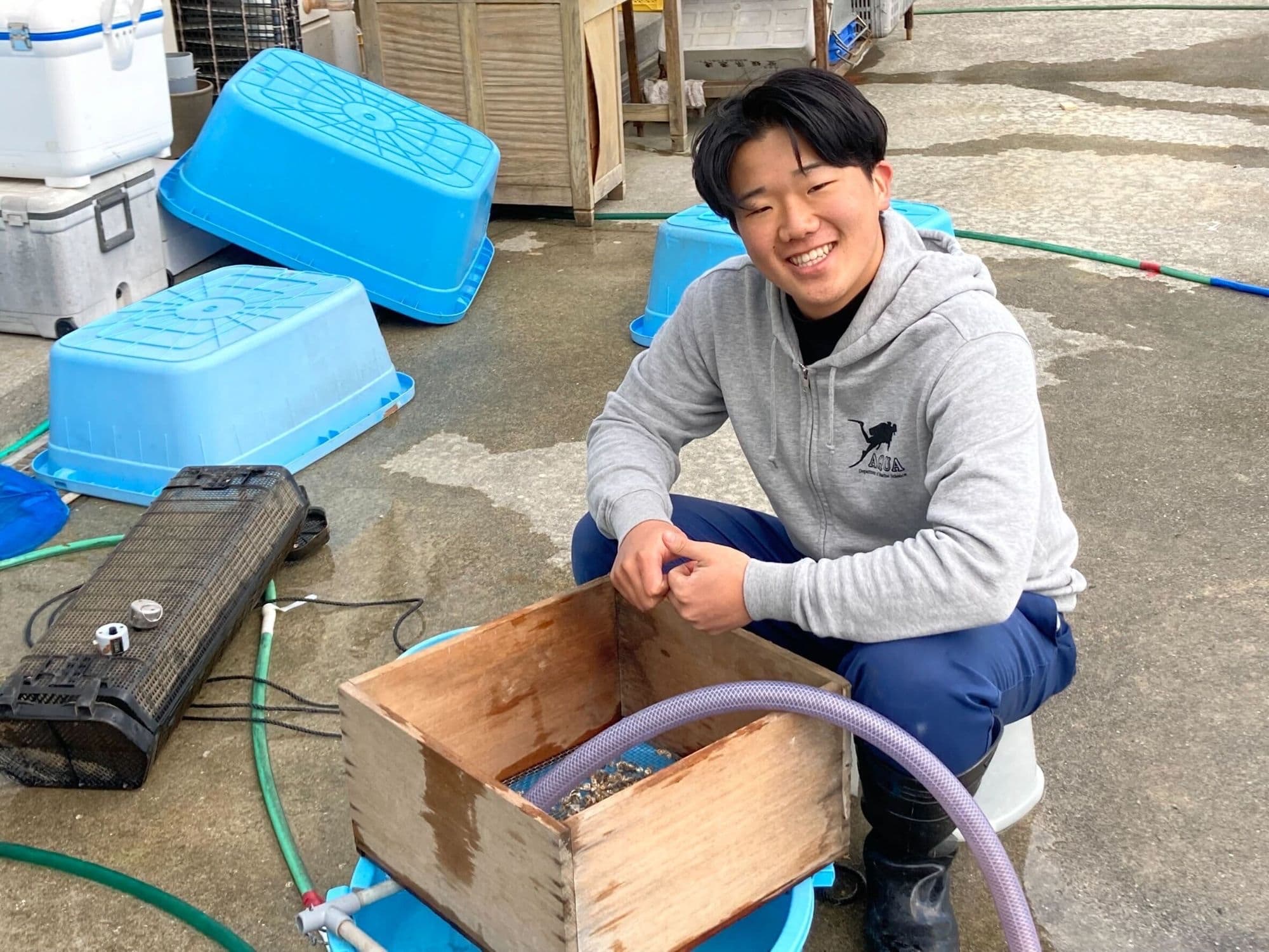漁場の作業場で、木箱のそばにしゃがみ、笑顔を見せる男性