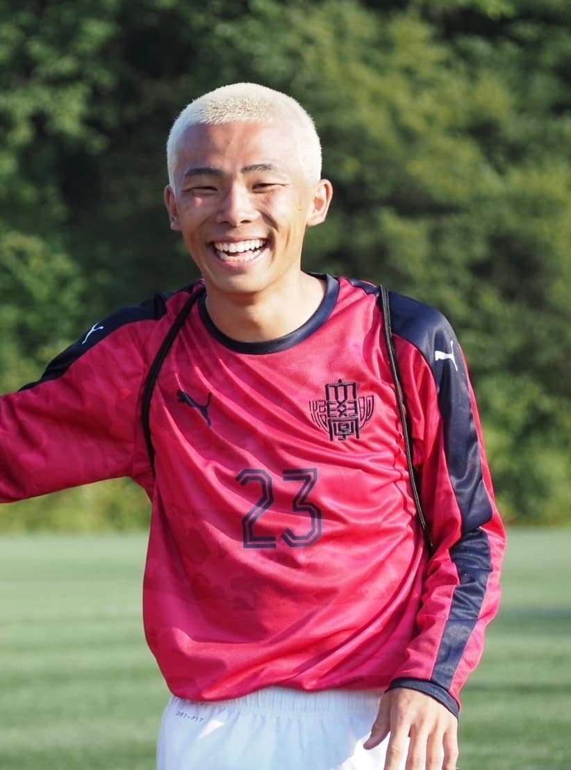 サッカーのユニフォーム姿で芝のグラウンドに立ち、笑顔を見せる男性